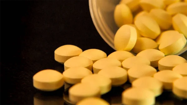 As pílulas amarelas espalhadas de uma garrafa transparente são espalhadas em uma mesa preta de vidro — Fotografia de Stock