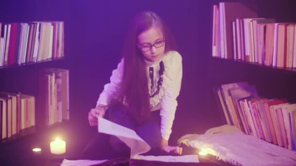 Uma menina abre um livro de contos de fadas na biblioteca, fumaça colorida gira ao redor, velas são acesas nas proximidades — Vídeo de Stock