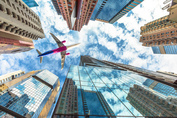 Airplane flying over modern skyscrapers on Hong Kong island. High-rise buildings in the blue dramatic sky. Concept of transport, travel and business. View from the ground level.