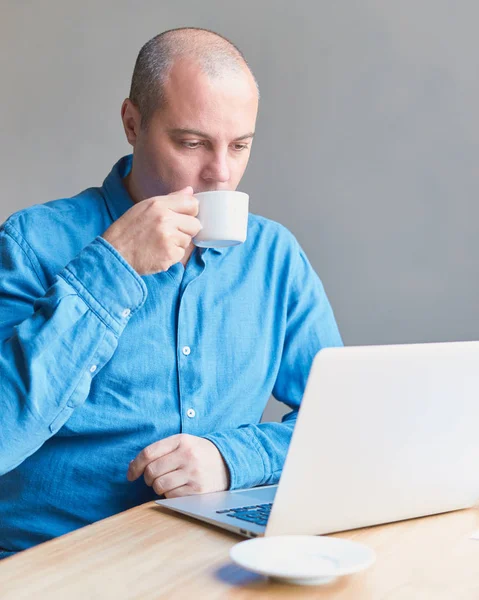 A handsome middle-aged man drinks coffee from mug and looks at screen of a computer, laptop. Man with casual wear in a blue shirt is sitting in office.