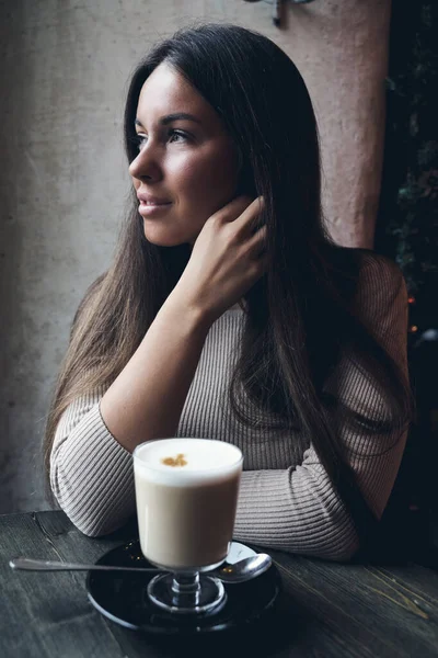 Beautiful girl sits in cafe in Christmas holidays, in background lights of garland. Brunette woman with long hair drinks cappuccino coffee, latte and looks out window, dark backdrop