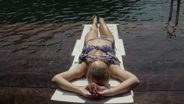 Frau liegt auf einer Sonnenliege mit Sonnenbrille und einem Boho-Seidentuch. Mädchen ruhen auf einem Flutholz-Unterwassersteg. Das Pflaster ist mit Wasser im See bedeckt.