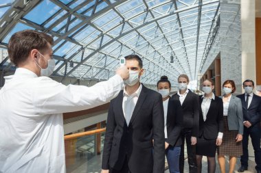 Koruyucu cerrahi maske takan doktor, kızılötesi alın termometresi (termometre tabancası) kullanarak bir kuyrukta bekleyen iş adamlarının vücut sıcaklığını kontrol ediyor.
