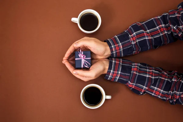 The male hands are closed on a brown background. The guy gives a gift to a girl. Mens hands next to two cups of coffee and with gift.