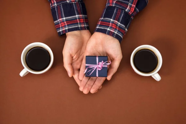 The male hands are closed on a brown background. The guy gives a gift to a girl. Mens hands next to two cups of coffee and with gift.
