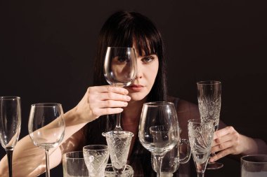 Siyah arka plandaki masada farklı şarap kadehleri olan genç bir kadın. Güneş ışığı ve gölgesi olan içecekler için bir sürü bardak..