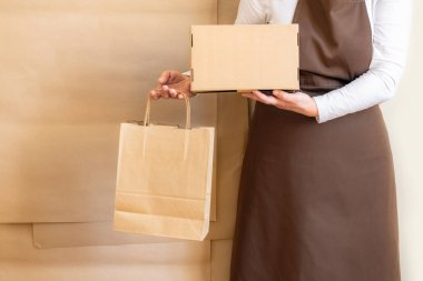 Paketçinin elleri bir Kese Kağıdı ve Kutu tutuyor. Kahverengi önlük giymiş işçiler Müşteri Paketleme Emri.