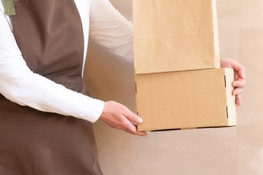 Paketçinin elleri bir Kese Kağıdı ve Kutu tutuyor. Kahverengi önlük giymiş işçiler Müşteri Paketleme Emri.