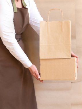 Paketçinin elleri bir Kese Kağıdı ve Kutu tutuyor. Kahverengi önlük giymiş işçiler Müşteri Paketleme Emri.