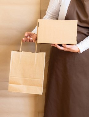 Paketçinin elleri bir Kese Kağıdı ve Kutu tutuyor. Kahverengi önlük giymiş işçiler Müşteri Paketleme Emri.
