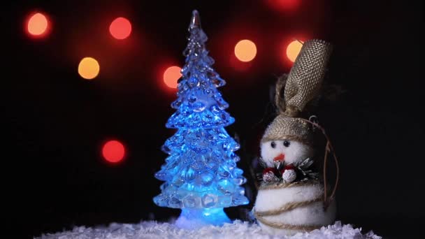Um boneco de neve dança em torno de uma árvore de Natal em um fundo de luzes coloridas — Vídeo de Stock