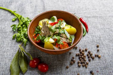 Kil tabakta Hashlama. Bileşenler: sığır pirzolası, patates, otlar, sarımsak. Tabak desenli gri bir tuval üzerinde duruyor. Etrafında kırmızı biber ve yeşil kabuklar, bezelyeler, domatesler, körfez yaprağı, maydanoz