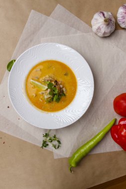 Karidesli peynir çorbası. Sarı çorba limon ve maydanozla süslenir. Beyaz yuvarlak bir tabak beyaz ve gri bir ambalaj kağıdının üzerinde yer alır. Biber, bezelye, domates, sarımsak, bitkilerle kaplıdır.. 