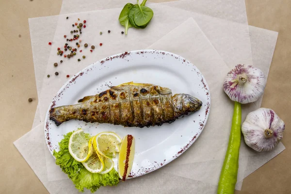 Trota alla griglia con limone e lattuga su un piatto bianco. Il piatto è su carta da imballaggio bianca e grigia, foderato con pepe, piselli, pomodori, aglio, erbe aromatiche — Foto Stock