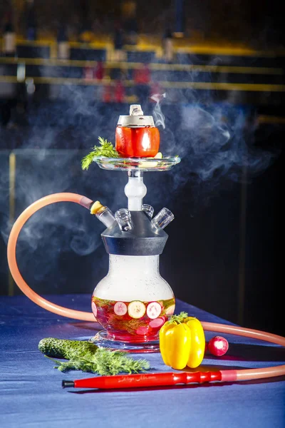 Hookah on vegetables. Hookah with a round transparent flask, which contains chopped vegetables: pepper, cucumber, radishes, herbs. Vegetables are filled with red liquor.  Lots of smoke. — Stock Photo, Image