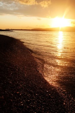 Gelendzhik Körfezi 'nde dramatik Altın Gün batımı. Güneş dağların ve bulutların ardında batar. Ön planda bir çakıl taşı plajı ve saman şemsiyeleri var.