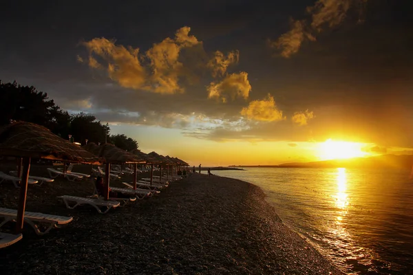 Dramatischer goldener Sonnenuntergang in der Gelendschik-Bucht. die Sonne geht hinter den Bergen und Wolken unter. Im Vordergrund ein Kieselstrand und Strohschirme — Stockfoto