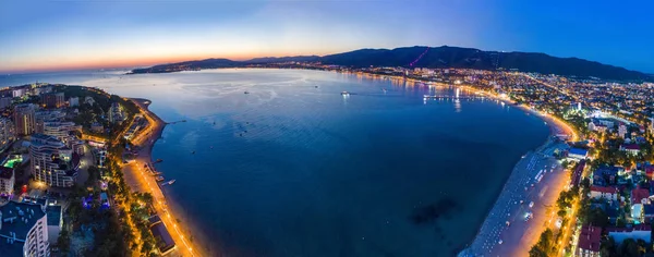 Gelendzhik sahil beldesinin akşam manzarası. Güneş battı, koyu mavi gökyüzü ve deniz. Visible Bay, plaj ve ışıklarda gezinti. Dağların siluetleri