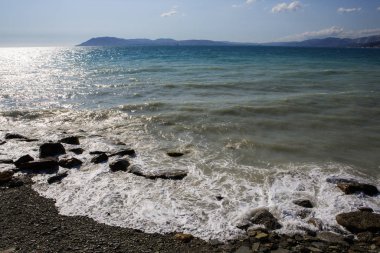 Karadeniz 'in Krasnodar bölgesindeki Kabardinka tatil beldesinde çakıl taşı plajı. Çakıl taşlı sahilde dalgalar ve sörf. Dağın ufukta Markoth bayırı görünüyor.. 