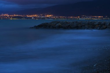 Akşamları deniz sörfü yapmak. Dalgalar çakıllı plajlarda ve kayalarda yuvarlanıyor. Arka planda, dağlar ve şehir ışıkları. 