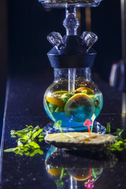 Nargile şişesi yuvarlak ve şeffaf, mavi likörle dolu. Dilimlenmiş portakal ve nane yaprakları likörde yüzer..
