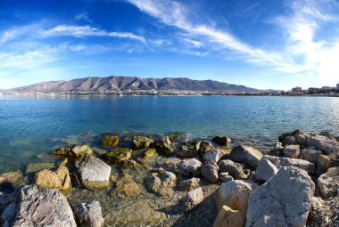 Autumn panorama of Gelendzhik Bay. Clear air, clear sky, and sharp clarity. In the foreground are rocks and a beach. In the background the Caucasus mountains