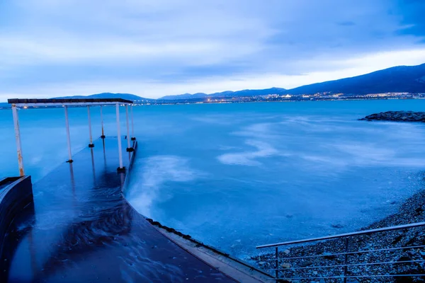 Praia de Gelendzhik de tarde em uma tempestade. As ondas a alta velocidade transformam-se num nevoeiro azul. Tons azuis, inverno. O cais vai para o mar. Praia de seixos . — Fotografia de Stock