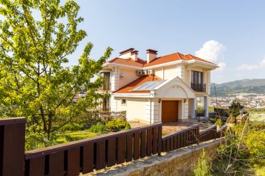 Kırmızı çatılı, iki katlı güzel bir kır evinin ön cephesi. Kulübe, kum taşından yapılmış taş sütunlar arasındaki ahşap bir çitin arkasında yer alıyor.. 