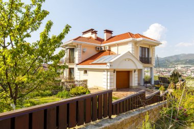 Kırmızı çatılı, iki katlı güzel bir kır evinin ön cephesi. Kulübe, kum taşından yapılmış taş sütunlar arasındaki ahşap bir çitin arkasında yer alıyor.. 