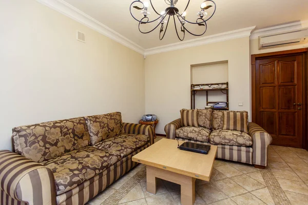 Bej renkli klasik oda. İki kahverengi kanepe, İskandinav ahşap sehpası ve yerde bej fayanslar. Dövülmüş zarif metal avize..