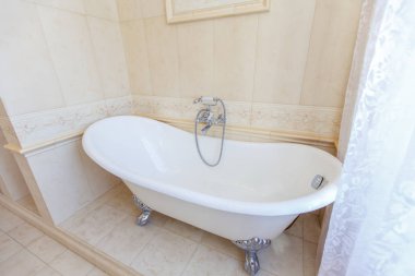 Klasik bej tonlu banyo. Kavisli banyo küveti. Büyük bej fayans. Yerde beyaz fayanslar var. Bükülmüş avize.