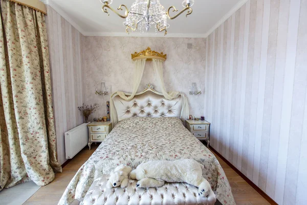 Elegant Royal white bedroom with a large double bed. White Carved back with a small canopy, white bedside tables with curved legs. Light striped Wallpaper. Lamps and chandelier \