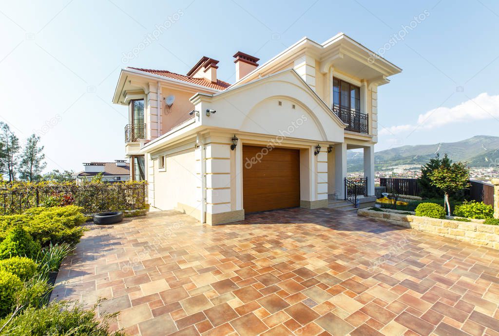 The facade of a two-story white classic cottage with a front garden. Carved entrance door, car gate, beige tiles.