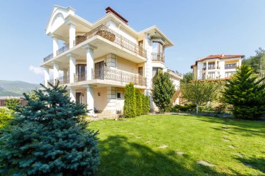İşlenmiş demir parmaklıkları ve sütunları olan üç katlı klasik bir kır evinin arka bahçesi. Kulübenin önünde yeşil bir çimen, ağaçlar ve boş bir havuz var.. 