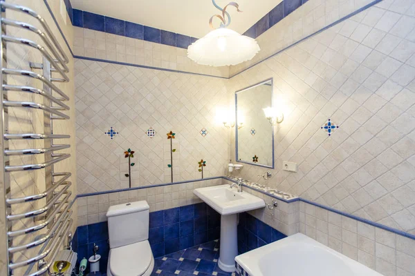 Classic rich beige bathroom with bathtub, toilet and sink with mirror. The walls are white tile with a pattern, the floor is blue tile.