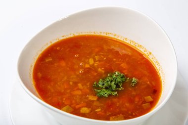 Dilli gulaş çorbası. Kırmızı çorbalı beyaz oval bir tabak beyaz arka planda duruyor.