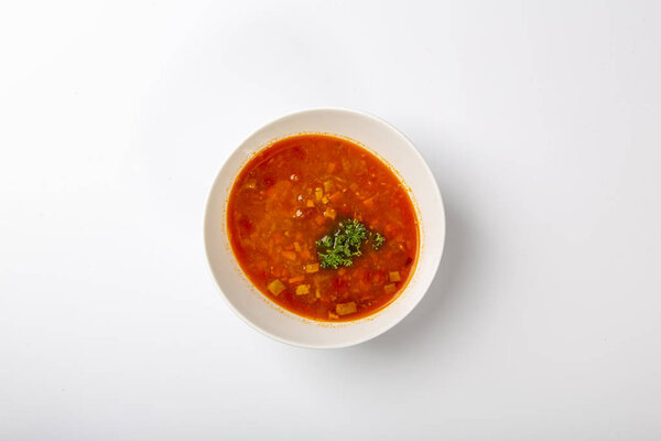 Говяжий гуляш с языком. Белая овальная тарелка с красным супом стоит на белом фоне. Вид сверху
