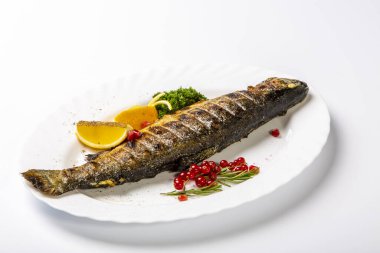 Limonlu ve otlu alabalık. Tabakta kafası ızgarada olan bir alabalık var. Limon, maydanoz ve frenk üzümünün yanında. Beyaz arkaplan üzerine plaka