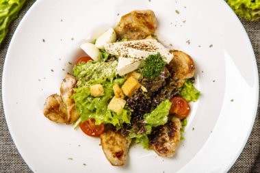 Sezar tavuklu ve peynirli. Yuvarlak beyaz tabakta servis ediliyor: kızarmış tavuk, peynir, domates, salata. Tabak gri bir tuval üzerinde