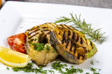 Izgarada mersin balığı bifteği. Kare tabakta ızgara mersin balığı bifteği var. Limon ve domatesle süslenmiş bitkilerle süslenmiş..