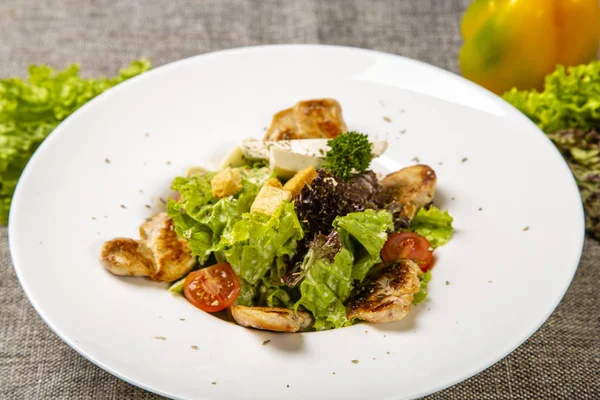 Sezar tavuklu ve peynirli. Yuvarlak beyaz tabakta servis ediliyor: kızarmış tavuk, peynir, domates, salata. Tabak gri bir tuval üzerinde