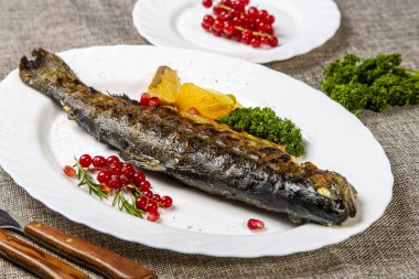 Limonlu ve otlu alabalık. Tabakta kafası ızgarada olan bir alabalık var. Limon, maydanoz ve frenk üzümünün yanında. Tabak gri bir tuvalde. Kırmızı biber, çatal, kaşık, ekmek sepetinin yanında.