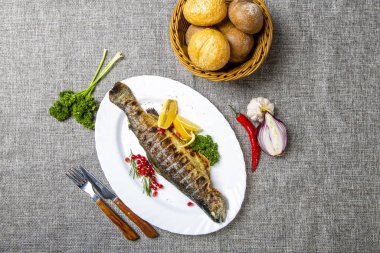 Limonlu ve otlu alabalık. Tabakta kafası ızgarada olan bir alabalık var. Limon, maydanoz ve frenk üzümünün yanında. Tabak gri bir tuvalde. Kırmızı biber, çatal, kaşık, ekmek sepetinin yanında.