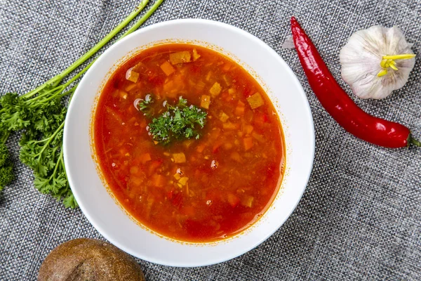 Rindergulaschsuppe mit Zunge. Ein weißer ovaler Teller mit roter Suppe steht auf einer grauen Leinwand. Daneben Paprika, Knoblauch, Kräuter und ein rundes Roggenbrot. Der Blick von oben — Stockfoto