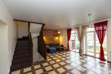 Spacious hall in the style of minimalism in the cottage. Comfortable blue sofa. Decorative red brick wall with lamps. Beige checkered floor. Wooden staircase to the second floor. 