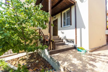 Veranda, merdivenler ve evin arka bahçesindeki kulübeye giriş. Girişteki tepe örtüsü. Ayakkabılar verandada. Ağaçlı arka bahçe. Güneşli bir gün.