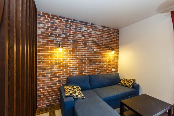 Um sofá azul com uma mesa de café na parte de trás da casa. Atrás do sofá é uma parede decorativa de tijolo vermelho velho com duas lâmpadas — Fotografia de Stock