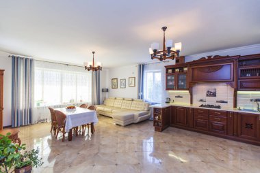 Büyük, parlak, güneşli bir salon. Odanın ortasında beyaz bir masa örtüsü ve dört hasır sandalyeli bir masa var. Beyaz deri kanepe. Klasik ahşap mutfak büfesi..