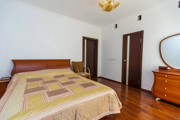 A large bedroom with a mahogany bed, wicker chairs, and an elegant dressing table with a semi-circular mirror. The bed has a beige blanket in squares. mahogany parquet — Stock Photo, Image