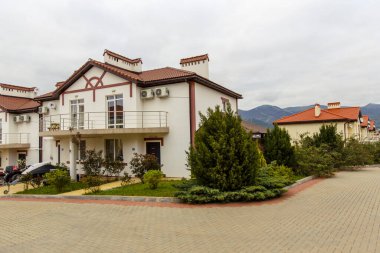 Baharda bulutlu havada iki katlı beyaz bir kulübe. Kırmızı çatılı beyaz ev, asfalt döşemeler, çiçekler. Ufuktaki dağlar